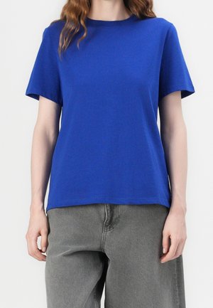 Femme portant un t-shirt uni bleu roi à manches courtes et un pantalon gris ample, debout sur un fond blanc.