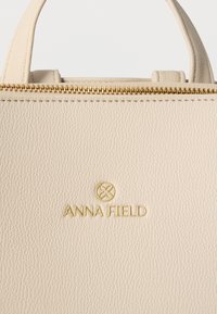 Τσάντα χειρός από δέρμα με υφή κρέμας, με χρυστό φερμουάρ και χρυσό λογότυπο «ANNA FIELD» στο κέντρο κάτω από το φερμουάρ, και μικρές λαβές στην κορυφή.