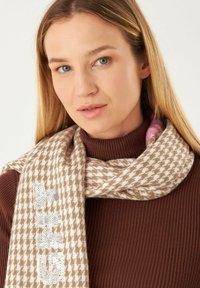 Bruine en crèmekleurige houndstooth sjaal met paillettenaccenten, gestructureerde stof, gedrapeerd om de hals, complementair aan een bruin coltrui.