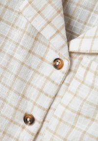 Camicia di lino con un motivo a quadri bianco e beige, con una texture morbida e due bottoni marroni al colletto e alla patta.