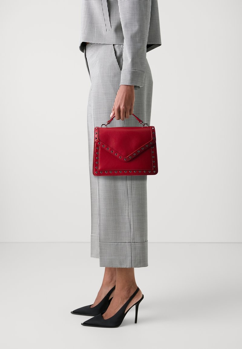 Personne portant un costume à carreaux noir et blanc, tenant un sac à main rouge clouté, chaussée de slingbacks noires à talons hauts, debout devant un fond uni.