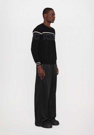 Un pull en tricot noir présente le mot 'ICEBERG' écrit en gris foncé, avec une bande horizontale blanche sur les épaules ; accompagné d'un pantalon noir ample et de chaussures noires.