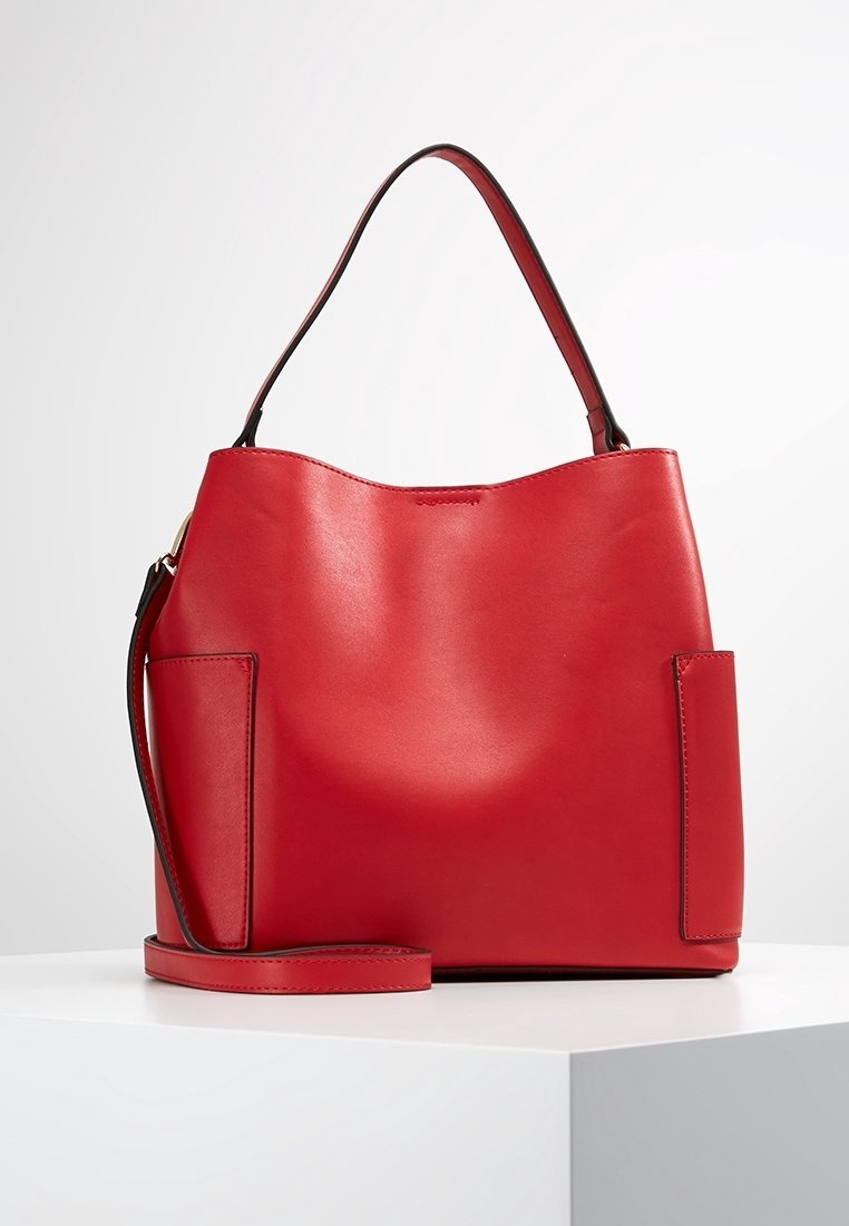 Červená kožená kbelík taška s hladkým povrchem, vyznačuje se jedním horním uchopovací rukojetí a bočními kapsami. Obsahuje odnímatelný ramenní popruh.