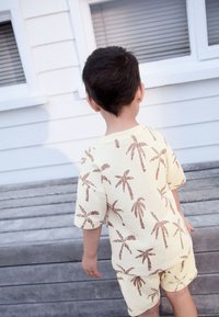 Žuti set od pamučne majice i kratkih hlača s motivom smeđeg palminog drveta. Opuštenog kroja, kratkih rukava, a hlače su do koljena, postavljeno na drvenoj pozadini.