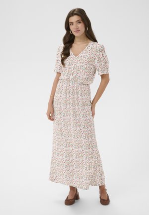 Femme aux longs cheveux bruns portant une robe longue maxi blanche à manches courtes à fleurs et des chaussures à talons carrés tressées marron, debout avec une main sur la taille.