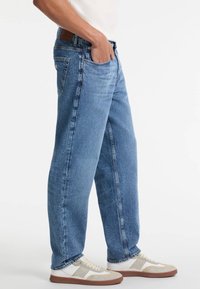 Jeans de mezclilla azul claro con un diseño de pierna recta, cinco bolsillos y un parche de cuero marrón en la cintura trasera, llevados con zapatillas blancas y grises.