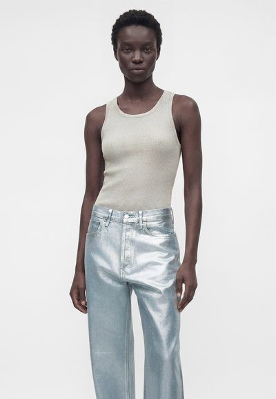 Γυναίκα που φοράει αμάνικη μπεζ ριγωτή μπλούζα και ψηλόμεσο μεταλλικό ασημί παντελόνι, στέκεται μπροστά σε λευκό φωτεινό φόντο.