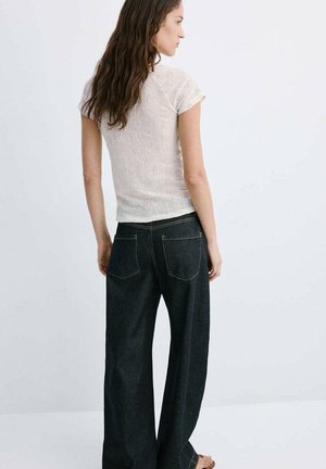 Femme aux longs cheveux ondulés portant un haut blanc texturé à manches courtes et un jean ample en denim foncé, debout devant un fond clair uni.