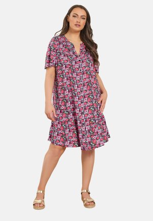 Kvinde stående med hånden i lommen iført en knælængde lyserød blomsterkjole med korte ærmer og beige sandaler.