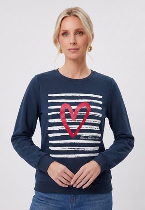 Femme blonde portant des boucles d'oreilles créoles en or et un sweat-shirt bleu marine avec des rayures blanches et un motif cœur rouge, mains jointes lâchement devant.