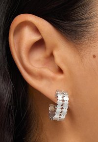 Boucles d'oreilles créoles argentées au design ondulé, composées de segments interconnectés et festonnés. Texture lisse avec une finition polie.