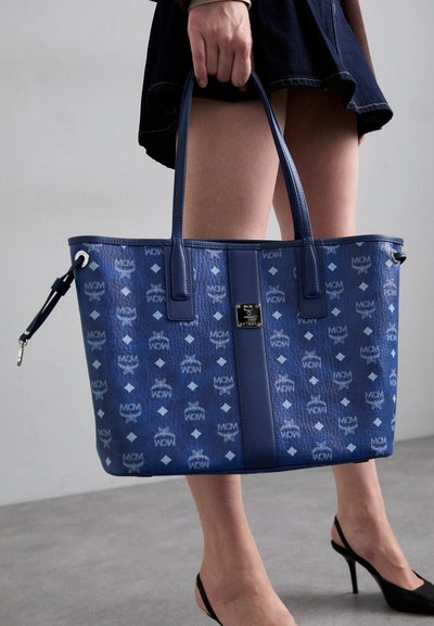 Geantă bleu tip tote cu o suprafață texturată, având un model repetat cu logo-ul MCM, completată de mânere din piele elegante și accente din metal argintiu.