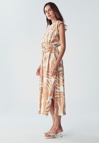 Maxi šaty v béžové barvě s bílými abstraktními vzory; mají krátké rukávy, elastický pas a boční rozparek. V kombinaci s bílými sandály na podpatku.