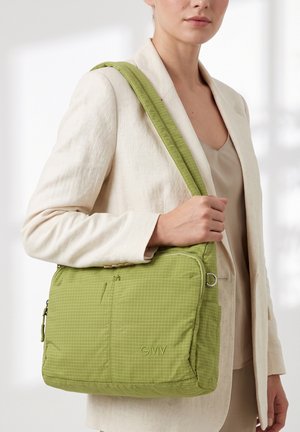 Vrouw met beige blazer houdt een groene geruite schoudertas vast met zakken met rits aan de voorkant en "GMV" logo.