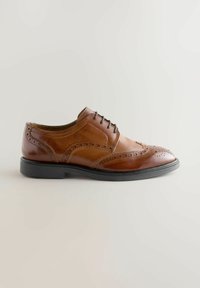 Zapato de vestir brogue de cuero marrón con suela y cordones negros, que presenta perforaciones decorativas y un diseño en punta en un fondo neutro.