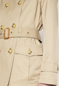 Beige trenčkot vyrobený z texturované látky. Obsahuje velké žluté knoflíky, pásek s obdéln�íkovou sponou a dvě přední kapsy.