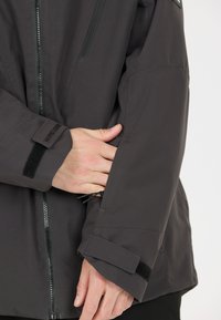 Donkergrijze jas met een ritssluiting aan de voorkant, verstelbare manchetten met klittenbandsluiting en een slanke, gestructureerde afwerking.