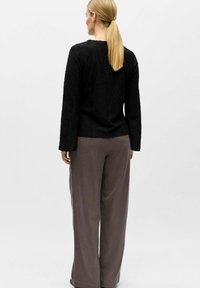 Vrouw met blond paardenstaart, gekleed in een zwart textuurachtig shirt met lange mouwen en losse grijze broek, staand en met haar rug naar de camera op een effen witte achtergrond.