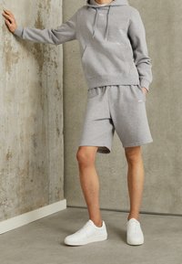Sudadera de algodón gris con bolsillo delantero y cordones, combinada con pantalones cortos grises a juego. Completo con zapatillas blancas sobre un fondo de concreto texturizado.