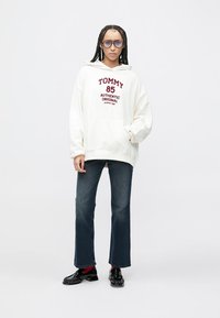 Κρεμ φούτερ με κουκούλα και εκτύπωση "TOMMY" και "85" σε μπορντό χρώμα. Συνδυασμένο με σκούρα μπλε καμπάνα τζιν και γυαλιστερά μαύρα παπούτσια.