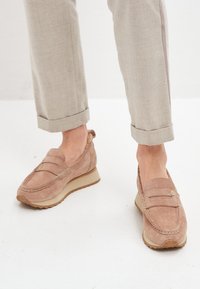 Mocasines de ante en tono tan con suela de cuña baja, que presentan costuras de mocasín y una tira decorativa en el empeine. Textura suave y forma redondeada.