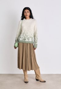 Pullover mit grünem, Farbverlauf-blumigem Muster, plissierter Midirock in Beige, beiger kniehoher Stiefel und eine kleine grüne Umhängetasche.