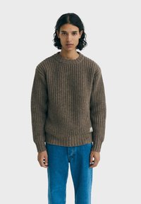 Pull en tricot marron avec une texture côtelée, col rond et manches longues, associé à un jean en denim bleu. Comprend un détail de petite étiquette.