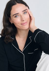 Haut de pyjama noir avec des bordures blanches, doté d'un col et d'une poche. Le tissu semble lisse et léger. Design minimaliste.