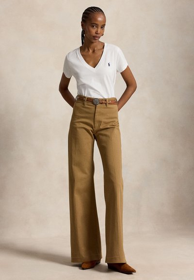 Femme portant un t-shirt blanc à col en V, un pantalon large taille haute beige, une ceinture marron avec une boucle décorative, et des chaussures marron pointues, debout les mains derrière.