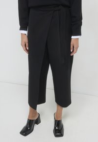 Pantaloni neri a wrapper realizzati in tessuto liscio, con una cintura da annodare in vita. Abbinati a mule neri lucidi con tacco a blocco.