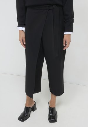 Schwarze Wickelhose aus glattem Stoff, mit einem Band in der Taille. Kombiniert mit glänzenden schwarzen Mules mit Blockabsatz.