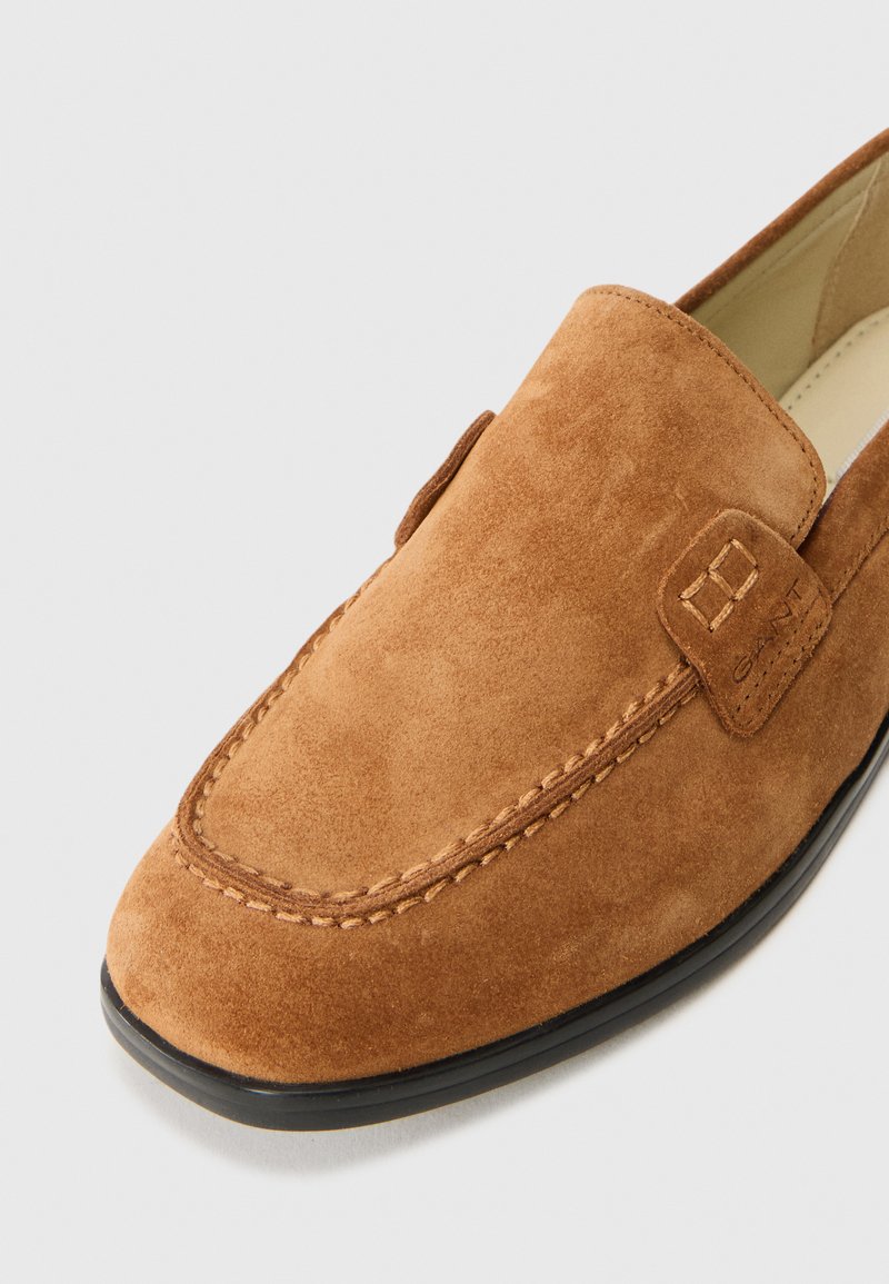 Brązowe zamszowe buty wsuwane z okrągłym noskiem, kontrastowym przeszyciem i małą skórzaną etykietą z logo po boku. Niska gumowa podeszwa.