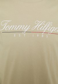 Brązowa bawełniana koszulka z białym, kursywowym napisem "Tommy Hilfiger" i "EST. 1985", z czerwonymi i czarnymi poziomymi liniami pod spodem.