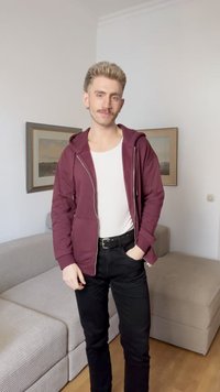 Sweatshirt com fecho marrom, com um bolso frontal, vestida sobre uma camisola branca. Jeans pretos com cinto. O fundo inclui um sofá de cor clara.