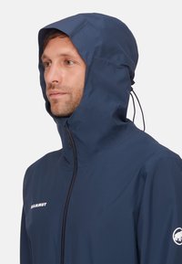 Giacca impermeabile blu navy con cappuccio, dotata di zip frontale e cordino elastico, logo del brand sul petto e sulla manica. Tessuto leggero.