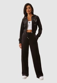 Schwarzer Kapuzenpullover mit Reißverschluss und Logo, kombiniert mit einem weißen Croptop und schwarzen Jogginghosen. Glatter Stoff, entspannte Passform und weiße Turnschuhe.