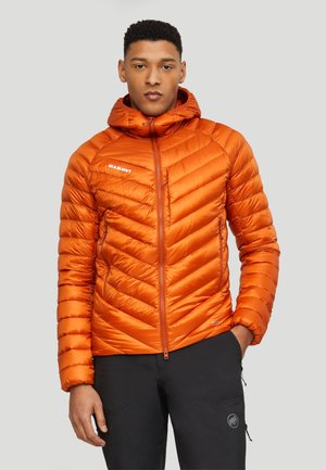 Mann trägt eine leuchtend orange gesteppt Kapuzenjacke und schwarze Hose mit Hand in der Tasche vor einfachem Hintergrund.