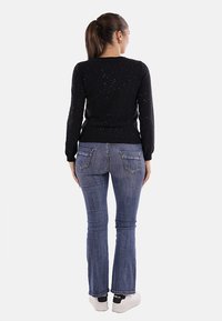 Donna di spalle che indossa un maglione a pois neri, jeans blu con tasche posteriori, sneakers bianche e orecchini a cerchio dorati, capelli raccolti in una coda di cavallo.