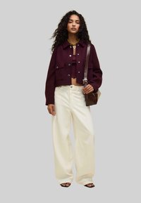 Burgundy cropped jacket with two front pockets, paired with a fitted top and white wide-leg pants; holding a small brown shoulder bag.