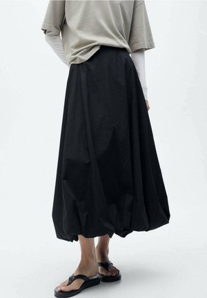 Personne portant une longue jupe noire bouffante, un haut beige à manches courtes par-dessus des manches longues blanches, et des sandales compensées noires.