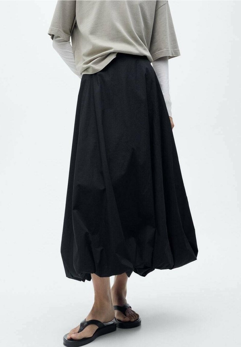 Personne portant une longue jupe noire bouffante, un haut beige à manches courtes par-dessus des manches longues blanches, et des sandales compensées noires.