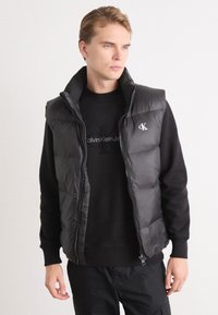 Gilet puffer nero con colletto alto e chiusura a zip. Presenta un logo Calvin Klein sul petto e una texture trapuntata su tutta la superficie.