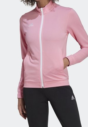 Kobieta w różowej kurtce na zamek z białym zamkiem i czarnych spodniach Adidas, z jedną ręką w kieszeni kurtki.