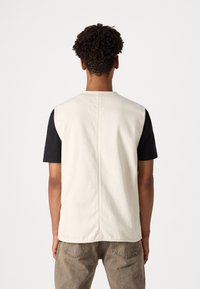 Ein ärmelloser cremefarbener Weste mit Rundhalsausschnitt, getragen über einem schwarzen T-Shirt, mit einer einfachen Rückennaht und kombiniert mit braunen, strukturierten Hosen.