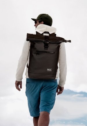 Brown waterproof roll-top backpack with a front zip pocket and adjustable straps, paired with a white sweater and blue shorts.