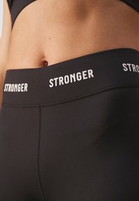 Leggings sportivi neri con vita alta e testo "STRONGER" in bianco. Il tessuto è liscio e presenta un design elasticizzato per il massimo comfort.