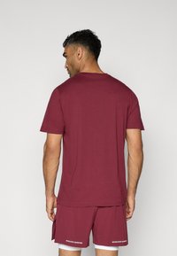 Maroons T-shirt med korte ærmer og rund halsudskæring, parret med matchende shorts med hvid tekstdetalje langs kanten.