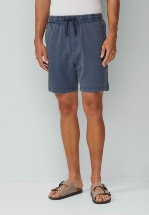 Dunkelblaue Shorts aus strukturiertem Stoff, mit elastischem Bund und Kordelzug. Tragen mit hellbraunen Sandalen, die eine dekorative Schleife haben.