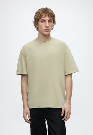 Junger Mann mit lockigem Haar, der ein lockeres beiges T-Shirt und dunkle Hosen trägt, steht vor einem schlichten weißen Hintergrund.