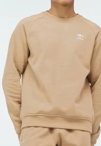 Sudadera beige de cuello redondo hecha de material suave, con un logotipo blanco de Adidas en el pecho y mangas raglán para un ajuste relajado.